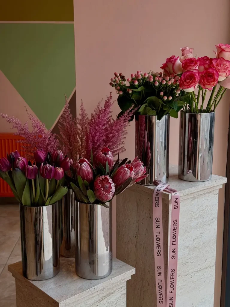 Üç adet gümüş renkli silindir vazo içerisinde ayrı ayrı sergilenen mor lale pembe protea pembe astilbe ve pembe gül çiçekleri.