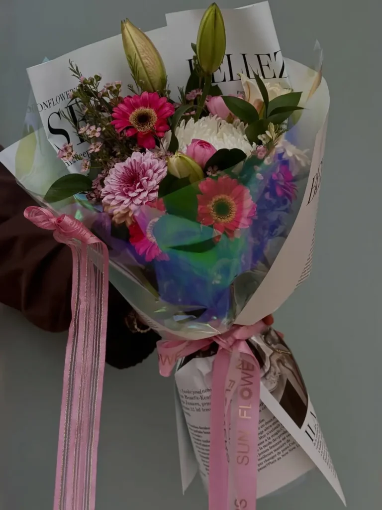 Parlak holografik jelatin ve gazete desenli ambalajlara sarılmış pembe kurdeleli beyaz krizantem ve taze pembe gerbera çiçekleri.