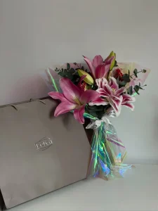 Şeffaf holografik ambalaj kağıdına sarılmış beyaz yazılı kurdeleli taze pembe lilyum çiçekleri ve yanındaki lüks gümüş renkli çikolata kutusu.