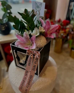 Pencereli siyah beyaz hediye çantası içinde pembe lilyum, beyaz bahar çiçekleri ve pembe kurdele.
