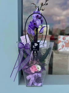 Siyah puantiyeli şeffaf çanta içerisinde mor kurdeleyle süslenmiş mor orkide beyaz papatya ve pembe karanfil çiçekleri bulunmaktadır.