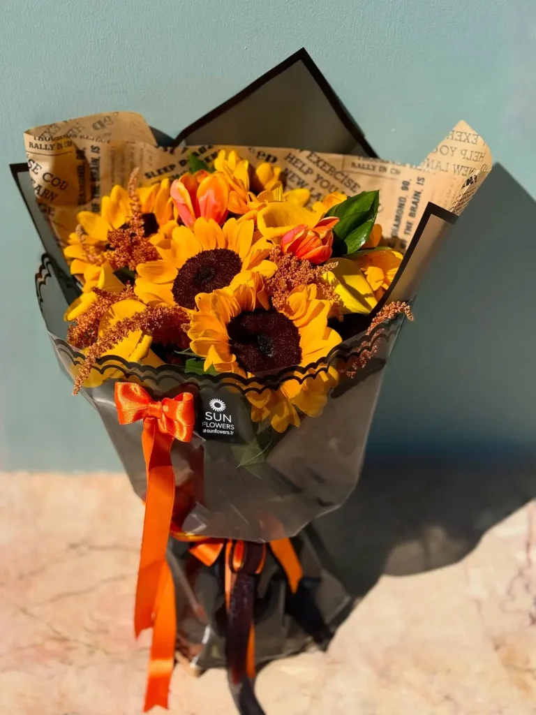 Siyah dantel kenarlı şeffaf ve gazete desenli ambalaj kağıtlarına sarılı turuncu kurdeleli turuncu laleler ve ayçiçekleri.