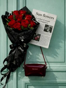 Siyah simli ambalaj kağıdına sarılı siyah kurdeleli kırmızı güller ve üzerinde Valentine's yazan bordo hediye çantası.