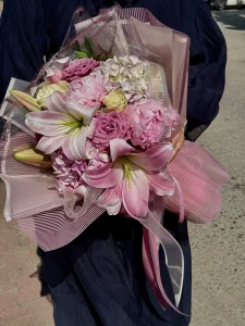 Üzerinde pleksi detaylı şerit bulunan toz pembe ambalajlı pembe lilyum ve şakayık buket tasarımı